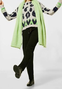 En ljusgrön scarf hänger över en grå tröja med svart leopardmönster och grönt geometriskt mönster, kombinerad med svarta byxor och svarta ankstövlar.