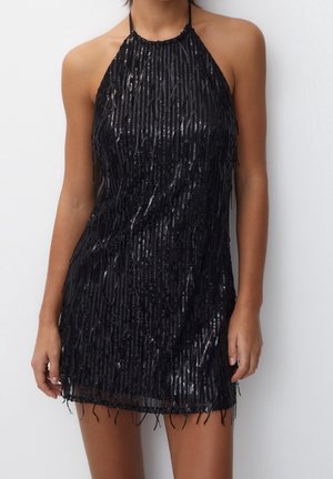 Femme portant une mini-robe noire sans manches ornée de sequins, avec des détails de franges verticales et un col halter, sur un fond blanc uni.