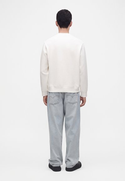 Homme vu de dos portant une chemise blanche à manches longues unie, un jean ample bleu clair et des chaussures noires sur un fond uni.