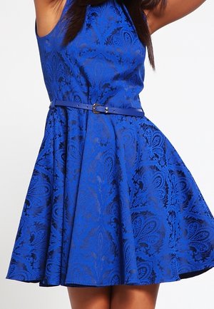Femme portant une robe bleue sans manches à motif floral avec une ceinture fine assortie, robe qui s'évase vers l'extérieur.