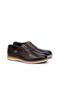 Zapatos de cuero marrón con un acabado suave, punta redondeada y suela blanca en contraste. Presentan cordones verdes y detalles de costura sutiles.