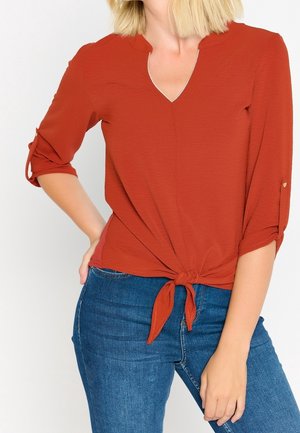 Femme portant un chemisier orange rouille à col en V, manches trois-quarts retroussées et noué devant, assorti à un jean bleu en denim.