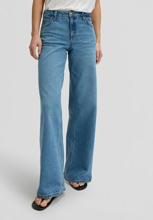 Kobieta ubrana w niebieskie, szerokie jeansy z wysokim stanem, białą koszulę włożoną do środka i czarne sandały z odkrytymi palcami na szarym tle.