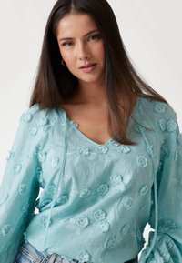 Lyseblå broderet bluse med blomsterretninger, en V-udskæring og lange ærmer med bindinger. Fremstillet af struktureret stof.
