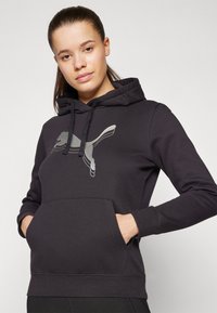 Juodas džemperis su kapišonu, frontinėje dalyje turi didelį atspindintį pilką Puma logotipą ir kengūros kišenę juosmens srityje.