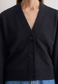 Cardigan noir en tissu doux, avec un col en V, fermeture à quatre boutons, ourlet côtelé et manches longues.