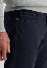 Mann in dunkelblauen Jeans und einem hellgrauen Langarmoberteil, mit entspannten Händen neben dem Oberschenkel.