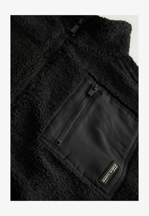 Czarna kamizelka z polaru o teksturowanej powierzchni, z zamkiem błyskawicznym w kieszeni i czarnym obszyciem. Podkreślona subtelną metką marki.