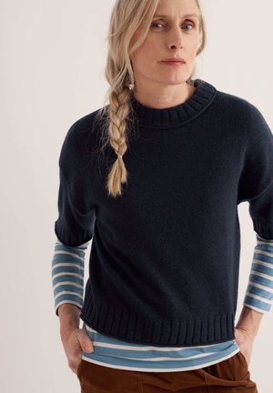Donna con capelli biondi intrecciati indossa un maglione blu navy sopra una camicia a righe blu e bianche, mani in tasca, rivolta in avanti contro uno sfondo semplice.