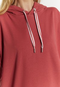 Felpa con cappuccio in un colore corallo tenue, caratterizzata da una tasca kangaroo frontale, tessuto texturizzato e lacci bianchi con punte in argento.