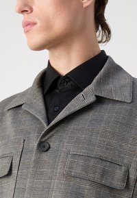 Grau karierte Jacke mit einem Kragen und zwei Fronttaschen. Verfügt über einen schwarzen Knopfverschluss und ein kontrastierendes schwarzes Hemd darunter.