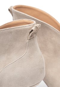 Béžové semišové členkové topánky s textúrovaným akcentom na päte, majú hladký vnútorný podšívku a zakrivený dizajn pozdĺž hornej časti.