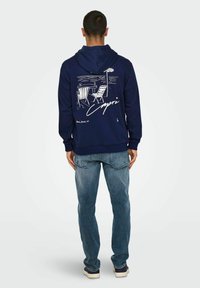 Marineblauer Kapuzenpullover mit einem Grafikdesign von zwei Strandst�ühlen und einem Tisch. Mit weißem Druckdetails und einer Kängurutasche. Kombiniert mit hellblauen Jeans.