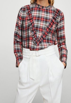 Vrouw die een rood-zwart geruit blouse met lange mouwen en ruchtdetails draagt, gecombineerd met wit, hoog getailleerd broek met riem.