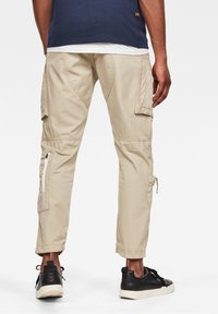 Beige cargo kalhoty s několika kapsami, zipovými detaily a nastavitelnými manžetami, kombinované s černými teniskami; látka má hladkou textura.