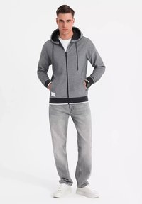 Sudadera gris con cremallera y capucha forrada en negro, con puños y cinturilla acanalados, combinada con jeans gris claro y zapatillas blancas.