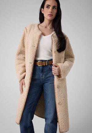 Femme portant un manteau beige texturé par-dessus un haut blanc côtelé et un jean bleu avec une ceinture marron, debout devant un fond gris uni.