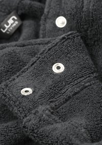 Musta froteekangas, jossa on näkyvä rakenne, varustettuna hopeisilla napilla ja tarralla. Kangas on pehmeä ja paksu, sopii käytettäväksi vaatetuksessa tai kodissa.