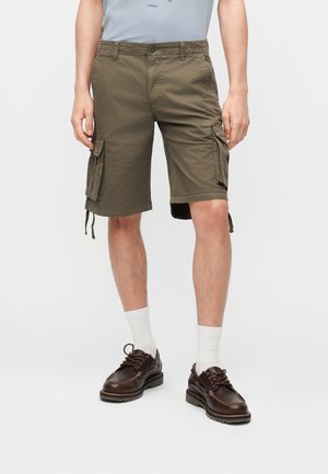 Personne portant un short cargo vert olive, des chaussettes blanches montantes et des chaussures en cuir marron foncé à lacets, debout devant un fond uni.