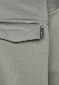 Primo piano di un tessuto grigio-verde con una tasca a patta e un piccolo etichetta nera con il logo Champion bianco e rosso sul bordo.