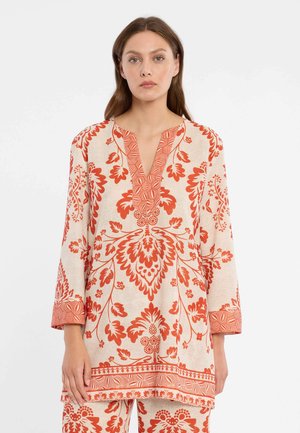 Femme portant une tunique beige et un pantalon ornés de motifs floraux et géométriques orange vif, debout devant un fond blanc uni.