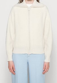 Kermanvärinen vetoketjullinen neuletakki, jossa on ribbineulottu pinta, leveä kaulus ja istuva helma. Yhdistetty vaaleansinisiin leveälahkeisiin housuihin.