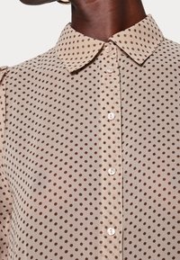 Camicia beige con pois marroni, dotata di colletto e chiusura con bottoni. Realizzata in un tessuto leggero e trasparente con una consistenza liscia.