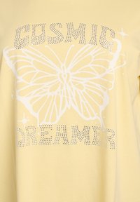 Camiseta de algodón amarilla con un gráfico de mariposa blanca y el texto "COSMIC DREAMER" adornado con tachuelas plateadas. Textura suave, ajuste relajado.