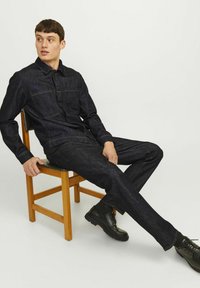 Chaqueta de mezclilla oscura y pantalones con detalles de costura en contraste. Textura suave, cierre con botones y dos bolsillos en el pecho. Zapatos negros sobre una silla de madera.