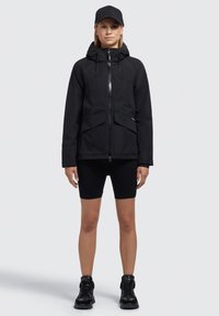 Giacca impermeabile nera con cappuccio, chiusura frontale con zip, due grandi tasche e design elegante. Indossata con pantaloni corti neri e robuste sneakers nere.
