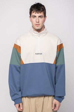 Pull-over à demi-zip couleur crème, mettant en avant des blocs de couleur vert, orange et bleu, avec un tissu texturé et un logo sur la poitrine.