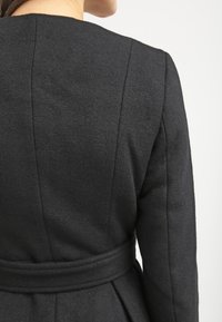Schwarzer Wollmantel mit einer figurbetonten Silhouette, rückwärtigen Nähten und einem passenden Gürtel in der Taille, mit einer strukturierten Oberfläche.