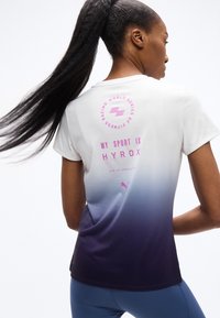 Γυναίκα με μακρύ αλογοουρά φορώντας αθλητική μπλούζα με ombre από λευκό σε μωβ και ροζ κείμενο «MY SPORT IS HYROX» στην πλάτη, και μπλε κολάν.