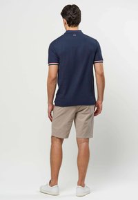 Man wearing navy polo with white, red, and blue striped sleeve cuffs, beige shorts, and white sneakers, standing facing away on white background.