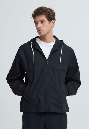 Junger Mann mit lockigem Haar trägt eine schwarze Kapuzenjacke mit Reißverschluss über einem weißen Hemd, die Hände in den Taschen, blickt seitlich vor einem einfarbigen Hintergrund.