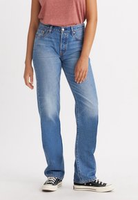 Vaqueros de mezclilla azul desgastados con un corte recto, cintura alta y un ligero efecto de desgaste. Combinados con zapatillas negras con suelas blancas.