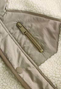 Jachetă din fleece bej, cu interior texturat crem, o secțiune din material contrastant și un buzunar lateral cu fermoar auriu.