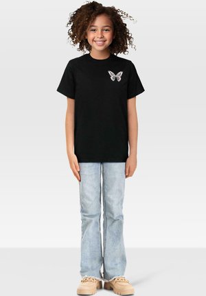 Svart bomulls t-skjorte med et sommerfugl grafikk på venstre bryst, kombinert med lyseblå flagrende jeans og beige sko.