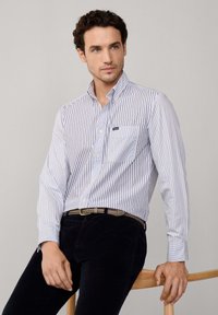 Camicia a righe bianca e blu in cotone, caratterizzata da una tasca sul petto. Abbinata a pantaloni scuri in cotone a coste e una cintura con fantasia.