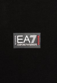 Černá tkanina s obdélníkovou etiketou, na které je v bílé a červené barvě vytištěno "EA7" a "EMPORIO ARMANI", spolu s ozdobným prošíváním.