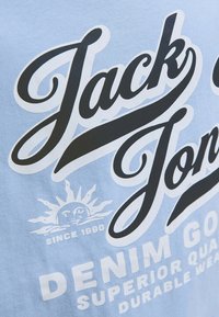Camiseta de algodón azul claro con texto en negrita en negro "Jack & Jones" y "Denim Goods" con acentos en blanco; gráfico del sol y detalle de "Desde 1990".
