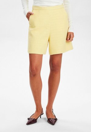 Yellow, textured shorts with a woven appearance. Features a concealed waistband and side pockets. Paired with burgundy pointy-toed shoes.
