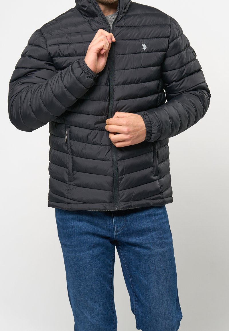 Svart pufferjacka med horisontell sömnad, dragkedjestängning, fast huva och elastiska mudd. Bärs med blå jeans.