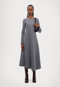 Gray, long-sleeved dress with a flared skirt and a smooth texture. Model holds a black bag, wears brown, knee-high boots. Simple and elegant design.