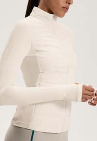 Lightweight beige jacket with a high collar, featuring a quilted texture on the body and smooth, fitted sleeves. No visible hardware.