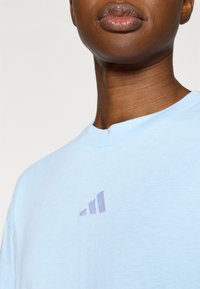 Ανοιχτό μπλε βαμβακερό μπλουζάκι με μικρό μωβ λογότυπο Adidas και στρογγυλή λαιμόκοψη. Το ύφασμα έχει απαλή, λεία υφή.