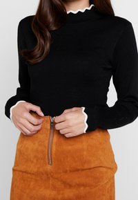 Svart ribbad långärmad topp med scallopmönstrad vit kant vid halsen och ärmsluten, ihopparad med en texturerad orange mockakjol med dragkedja fram.