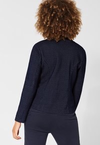 Námořnická modrá texturovaná bunda s vystouplým tečkovaným vzorem, dlouhými rukávy a přiléhavým střihem. Viditelný šev na zadní straně a žádné další kovové prvky.