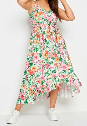 Frau trägt ein ärmelloses, weißes High-Low-Kleid mit pinken, orangefarbenen und grünen Blumen, kombiniert mit weißen Sneakers, posiert mit erhobenem Arm.