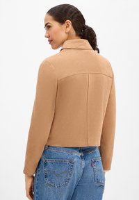 Giacca corta in camoscio color cuoio con colletto, caratterizzata da una texture liscia e dettaglio con cucitura posteriore, abbinata a jeans blu in denim.